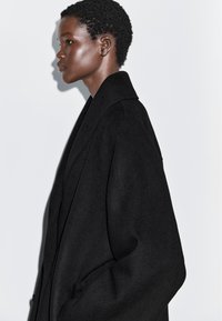 Profiel van een persoon met kort, krullend haar die een oversized zwarte jas draagt, tegen een effen lichte achtergrond.