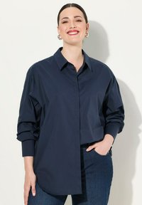 Camisa de gran tamaño de color azul marino con botones y cuello, de mangas largas y textura sutil. Combinada con jeans de mezclilla oscuros, que cuentan con bolsillos frontales.