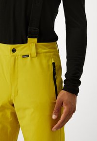 Pantaloni de schi galbeni cu suspensi ajustabili negri, buzunar frontal cu fermoar, textură netedă și închidere cu buton la talie.