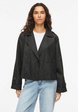 Femme aux longs cheveux bouclés portant une veste noire courte à double boutonnage sur une chemise blanche et un jean bleu clair sur fond uni.