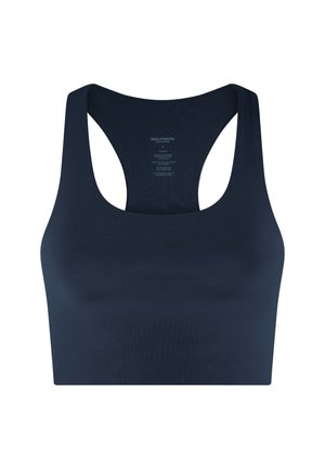 Fiatal nő göndör szőke hajjal, tengerészkék sportmelltartóban, fekete tréningnadrágban és fehér tornacipőben, egyszerű háttér előtt áll.