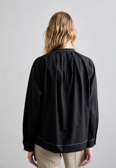 Blusa negra con tejido fruncido en la parte posterior, con detalles de costura blanca a lo largo del dobladillo y los puños. Material suave y liviano.