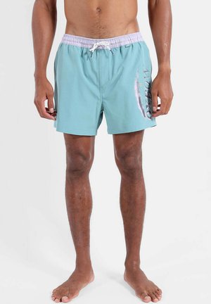 Homme portant un short de bain turquoise avec une ceinture blanche et un cordon blanc, debout pieds nus sur un fond uni.