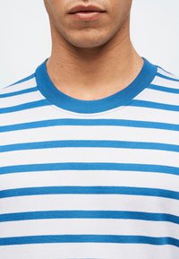 Άνδρας που φοράει λευκό μπλουζάκι με οριζόντιες μπλε ρίγες και μπλε λαιμόκοψη, εμφανιζόμενος από τον λαιμό μέχρι το στήθος.