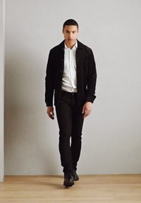 Hombre vestido con una chaqueta de pana negra sobre una camisa blanca, pantalones negros y zapatos negros, caminando sobre un suelo de madera frente a una pared gris lisa.