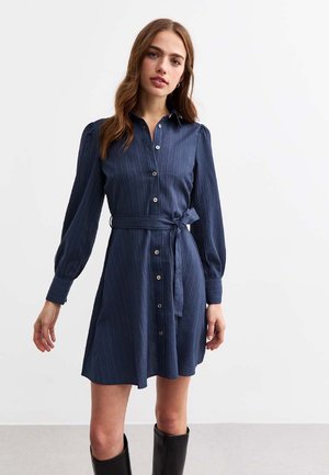 Navy knoopjurk met lange opgeblazen mouwen, een taille specifiek en een bijpassende strikceintuur. Verticale strepen en een subtiele glans door de stof.