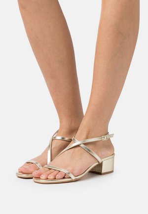Sandales à lanières métalliques dorées avec un talon bloc, dotées de fines lanières autour de la cheville et d'une semelle plate, présentant un design épuré et minimaliste.