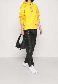 Geel hoodie met vo zakken aan de voorkant, zwarte leren broek en een kleine zwarte schoudertas. Witte sneakers met oranje accenten.