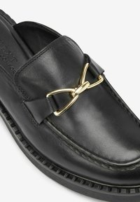 Mocassin en cuir noir avec un détail en métal doré en forme de mors sur le dessus et une semelle en caoutchouc noire texturée.