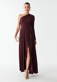 Bordeaux, een schouderjurk met lange mouw, aansluitend lijfje, vloeiende rok, zijopening en geplooide details. Gouden accenten met bandjes sandalen.