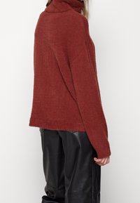 Hnědý rolák s volným střihem, vyrobený z měkkého, texturovaného materiálu. Má dlouhé rukávy a mírně zkrácený lem. Kombinuje se s černými kalhotami.