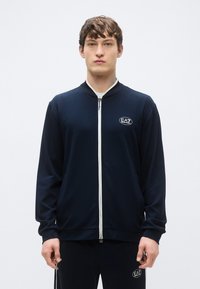 Giacca navy con zip realizzata in tessuto liscio, con colletto e zip bianchi, con un piccolo logo EA7 sul petto.