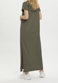 Olivegrünes Maxikleid mit kurzen Ärmeln, seitlichen Schlitzen und einem lockeren Schnitt. Kombiniert mit weißen Sportschuhen.
