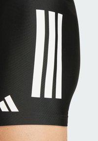 Pantaloni sportivi neri con tre audaci strisce bianche sui lati e una texture liscia, progettati per comfort e flessibilità.