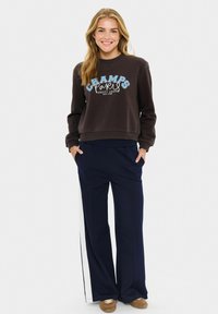 Felpa marrone con la scritta "CHAMPS", abbinata a pantaloni a gamba larga blu navy con strisce bianche sui lati. Il modello è in piedi con le mani nelle tasche.
