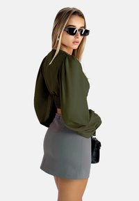 Olivgrünes kurz geschnittenes Oberteil mit Puffärmeln, kombiniert mit einem grauen Miniröckchen. Die Accessoires umfassen schwarze Sonnenbrillen und eine kleine schwarze Tasche.