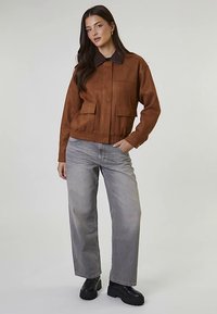 Brązowa zamszowa kurtka z czarnym kołnierzem, z dwoma przednimi kieszeniami i ściągaczem w talii, zestawiona z jasnoszarymi jeansami o szerokich nogawkach i czarnymi butami.