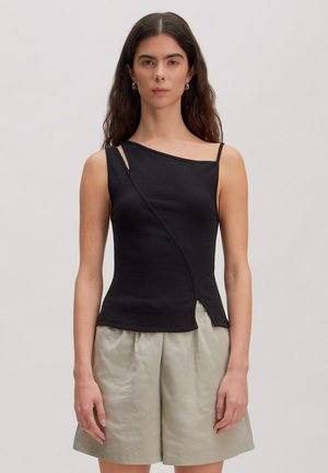 Frau mit langen dunklen Haaren, die ein ärmelloses asymmetrisches schwarzes Oberteil und lockere beige Shorts trägt, steht vor einem schlichten Hintergrund.
