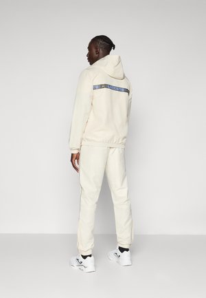Survêtements couleur crème avec une capuche, arborant une étiquette "LACOSTE" bleue et noire au dos. Associé à des chaussures de sport blanches.