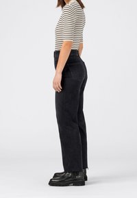 Frau in schwarzen, hoch taillierten, geraden Jeans, schwarzen klobigen Stiefeln und einem weißen Oberteil mit halben Ärmeln und schwarzen horizontalen Streifen, die seitlich steht.