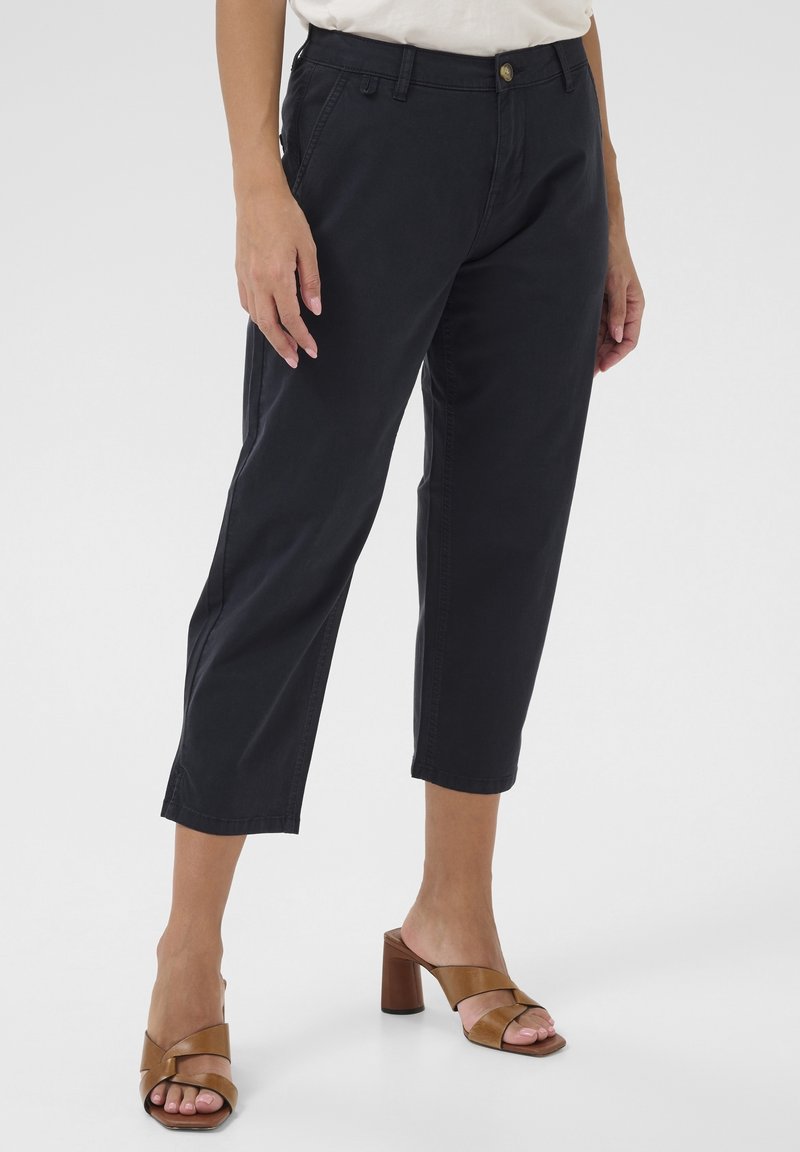 Pantalons noirs coupés en coton robuste, présentant une coupe classique, une fermeture à bouton et des poches latérales. Associés à des sandales à talons blocs marron.
