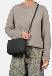 Bolso crossbody de cuero negro con un acabado suave, forma rectangular y correa ajustable, sostenido por una persona que viste un suéter beige.