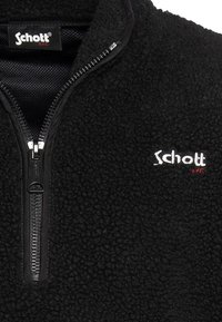 Czarna bluza polarowa z krótkim zamkiem i wysokim kołnierzem. Posiada biały napis "Schott" z czerwonym akcentem, wykonana z miękkiego, teksturowanego materiału.
