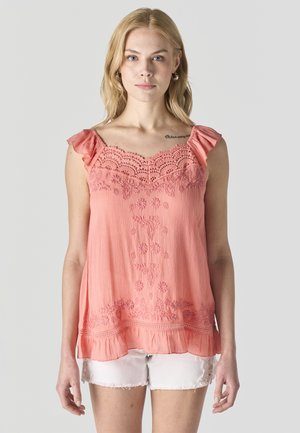 Joven mujer con blusa sin mangas coral rosa bordada y pantalones cortos blancos con encaje, de pie contra un fondo liso.