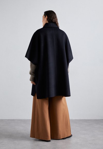 Cappotto di lana nera con un ampio colletto, vestibilità oversize e fessure laterali, abbinato a pantaloni larghi marroni. Il tessuto sembra morbido e texturizzato.