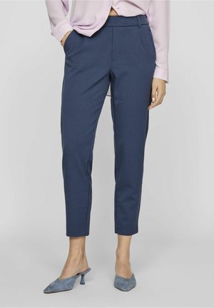 Pantalones azules marino a medida con corte slim, tejido estructurado, bolsillos delanteros y tobillos estrechos. Combinados con blusa rosa claro y tacones azules.