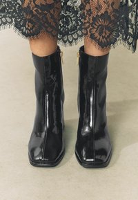 Black patent leather ankle boots with a square toe, featuring gold zipper accents on the sides, paired with a black lace skirt.