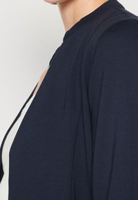 Granatowa bluzka o gładkiej fakturze, z wysokim kołnierzem i wycięciem po bokach. Materiał wydaje się lekki i dopasowany.