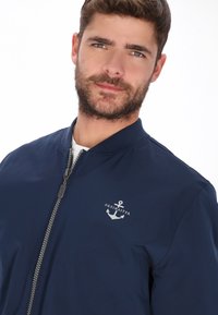 Marineblå bomberjakke med lynlås foran, ribbet krave og et sølvforankret logo på venstre bryst. Glat tekstur, letvægtsmateriale.
