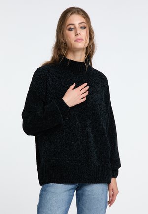 Junge Frau mit langen braunen Haaren, die einen schwarzen Strickpullover und hellblaue Jeans trägt und mit einer Hand ihre Brust berührt.