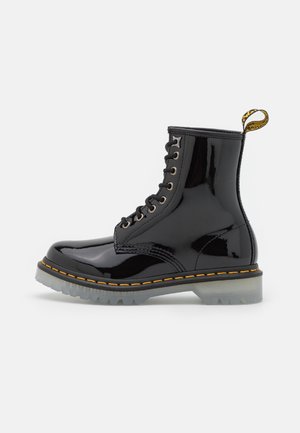 Bottines à lacets noires brillantes avec semelle transparente, couture jaune et une languette jaune et noire au talon.