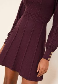 Gebreide jurk in diep bordeauxrood, met kabelgebreide patronen, een getailleerde taille en lange mouwen met gouden knoopaccenten bij de manchetten.