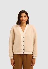 Cardigan in pile beige con scollatura a V, chiusura con bottoni e tasche laterali. Presenta una texture morbida e polsini a coste, indossato sopra pantaloni marroni.