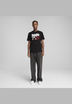 Black cotton t-shirt features bold white and red graphic print with "PUMA" and "1948." Model wears loose gray jeans and white sneakers.