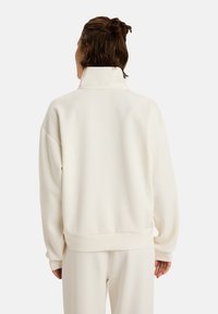 Pullover crème, réalisé en tissu doux, avec un col montant et des épaules tombantes. Il présente un ourlet et des poignets côtelés, au design minimal.