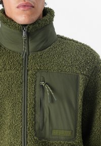 Chaqueta de forro polar verde oliva con cuello alto, cuerpo texturizado y panel liso. Cuenta con un bolsillo en el pecho con cremallera y cordones ajustables.