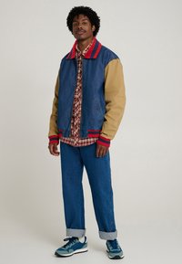 Denim bomber jacket with tan sleeves, red ribbed collar, and zip closure, layered over a red plaid shirt, paired with rolled blue jeans and sneakers.