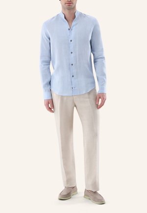 Mann trägt hellblaues, langärmeliges Hemd mit Knöpfen, beige Hose und beige Schlupfschuhe und steht vor einfachem Hintergrund.