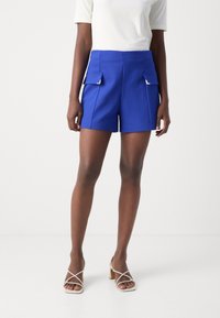 Blue shorts with two front pockets, tailored design, and smooth texture. Worn with a white shirt and light-colored heeled sandals.