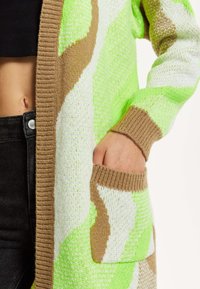 Knit cardigan featuring a wave pattern in neon green, cream, and brown. Ribbed edges with two front pockets. Soft texture.