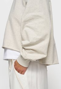 Felpa grigia chiara a maniche corte con maniche larghe e polsini a costine. Presenta un ricamo "10" sulla manica. Indossata sopra una camicia bianca.