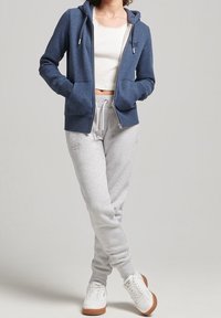 Sudadera con cremallera en color marino con un acabado texturizado, combinada con pantalones de chándal gris claro y zapatillas blancas, que presenta un corte casual y bolsillos laterales.