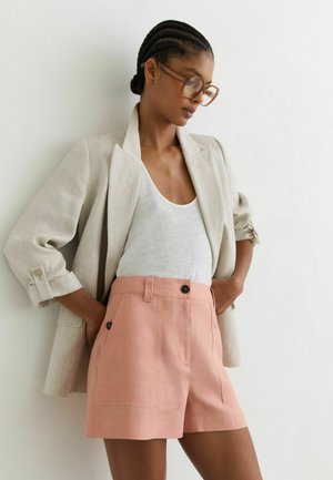 Vrouw met gevlochten haar die grote bril draagt, beige blazer over een witte top en high-waisted roze short, staand tegen een witte muur.