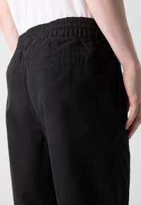 Personne portant un pantalon noir à taille élastique avec une seule poche passepoilée au dos, vue de côté et légèrement par derrière.
