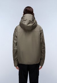 Persoon in een losse olijfgroene capuchonjack en zwarte broek, die met de rug naar de camera staat tegen een effen lichtgrijze achtergrond.