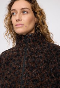 Braune Fleecejacke mit schwarzem Leopardenmuster, hohem Kragen und Reißverschluss vorne. Die Textur wirkt weich und plüschig mit kontrastierenden Farben.